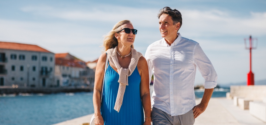 A smiling middle-aged man and woman walk side-by-side along a sunny, coastal boardwalk with a town and ocean in the background.